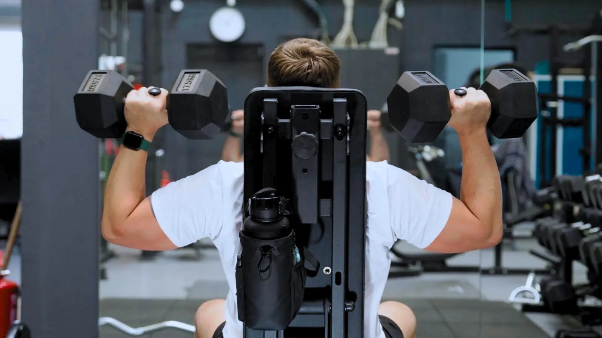 Bolsa magnética para gimnasio con portabotella colocada en máquina de entrenamiento mientras atleta levanta mancuernas.